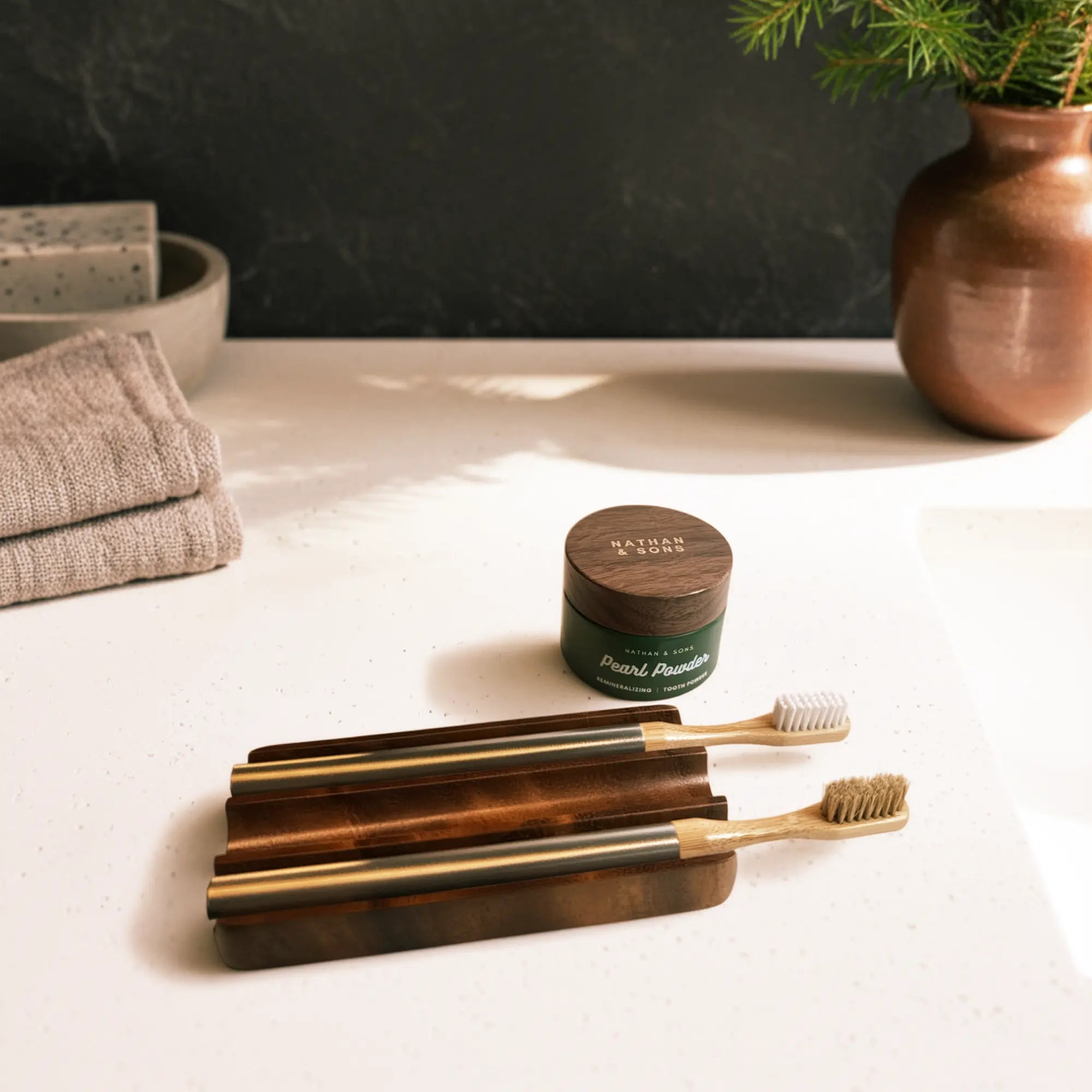Bathroom countertop with two bamboo toothbrushes featuring sleek aluminum handles resting on a wooden holder. One toothbrush has a natural boar’s hair bristle head, while the other features soft castor oil bristles resting beside a jar of Nathan & Sons Pearl Powder Remineralizing Tooth Powder.