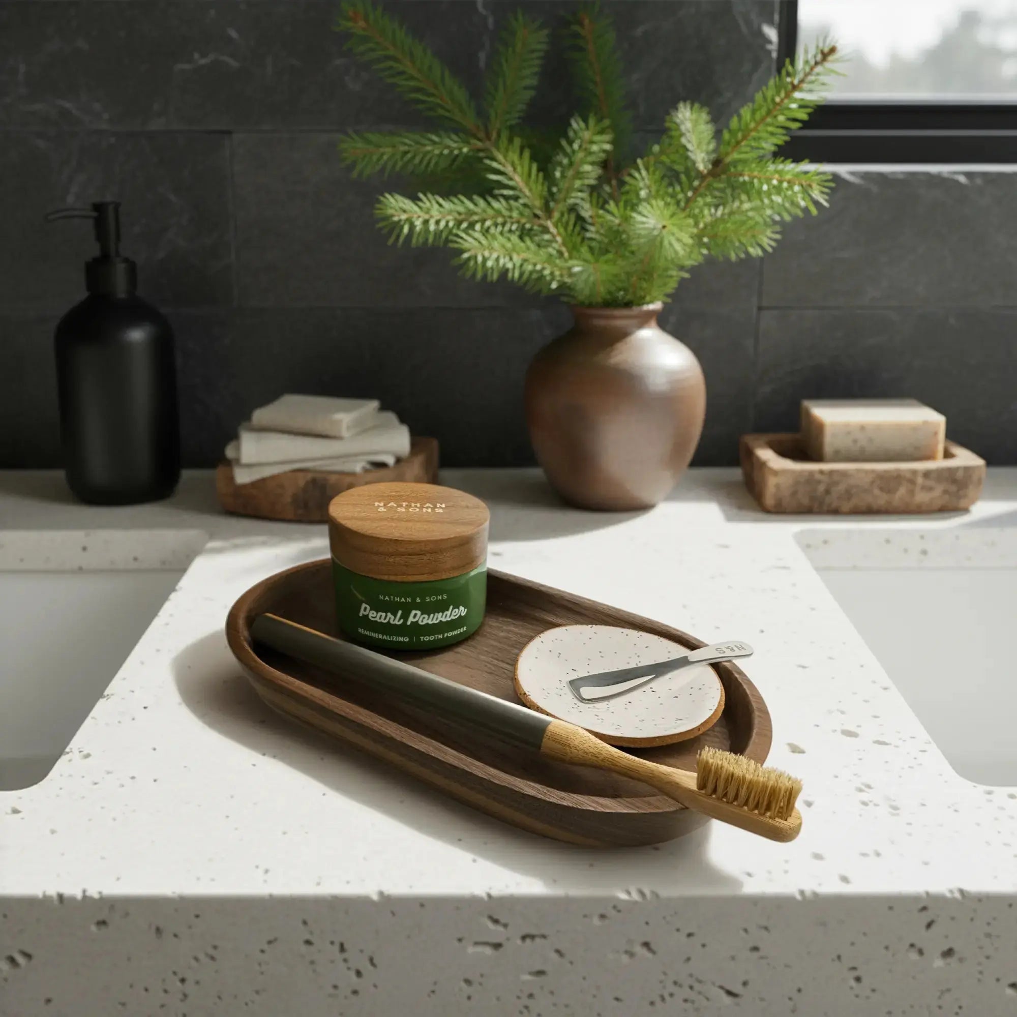 Bathroom countertop with a wooden tray holding a Nathan and Sons Boar's Hair Toothbrush Kit featuring boar's hair bristles, a bamboo neck and an aluminum handle; a Nathan and sons Speckled White Handmade Gum dish, and a jar of Nathan & Sons Pearl Powder Remineralizing Tooth Powder.