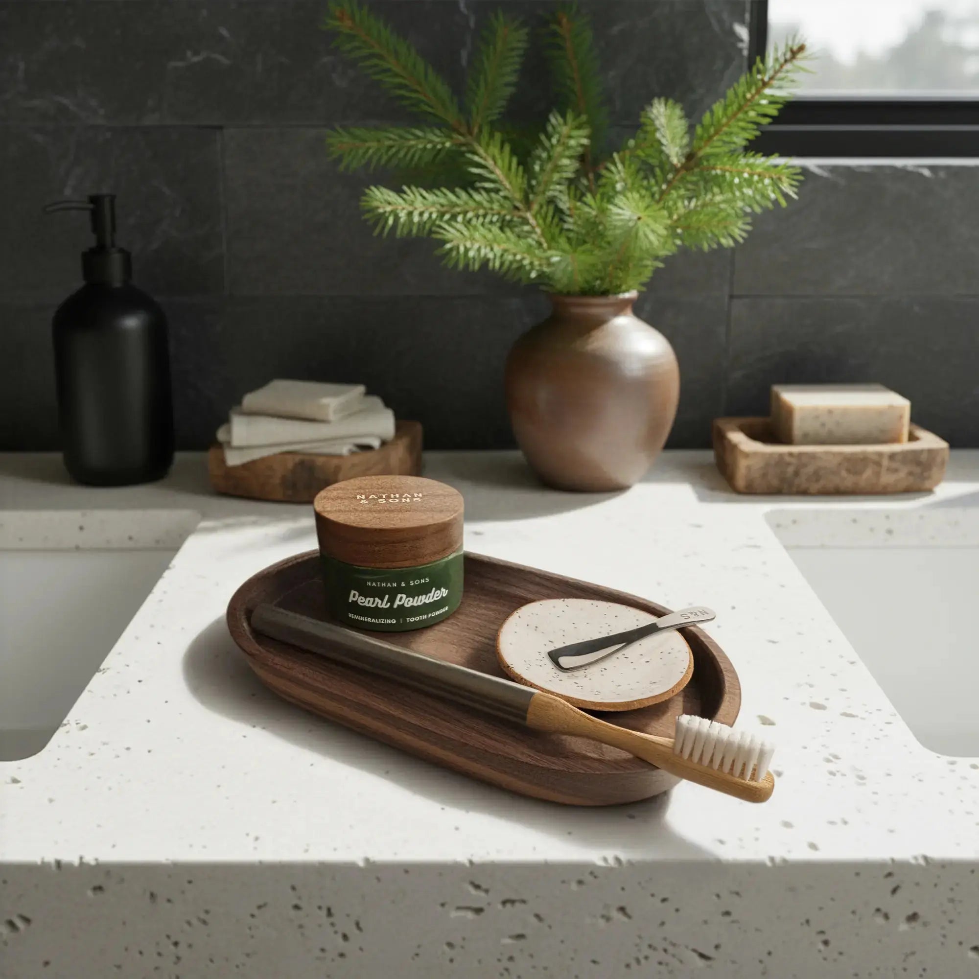 Bathroom countertop with a wooden tray holding a Nathan and Sons Castor Oil Toothbrush Kit featuring castor oil bristles, a bamboo neck and an aluminum handle; a Nathan and sons Speckled White Handmade Gum dish, and a jar of Nathan & Sons Pearl Powder Remineralizing Tooth Powder.