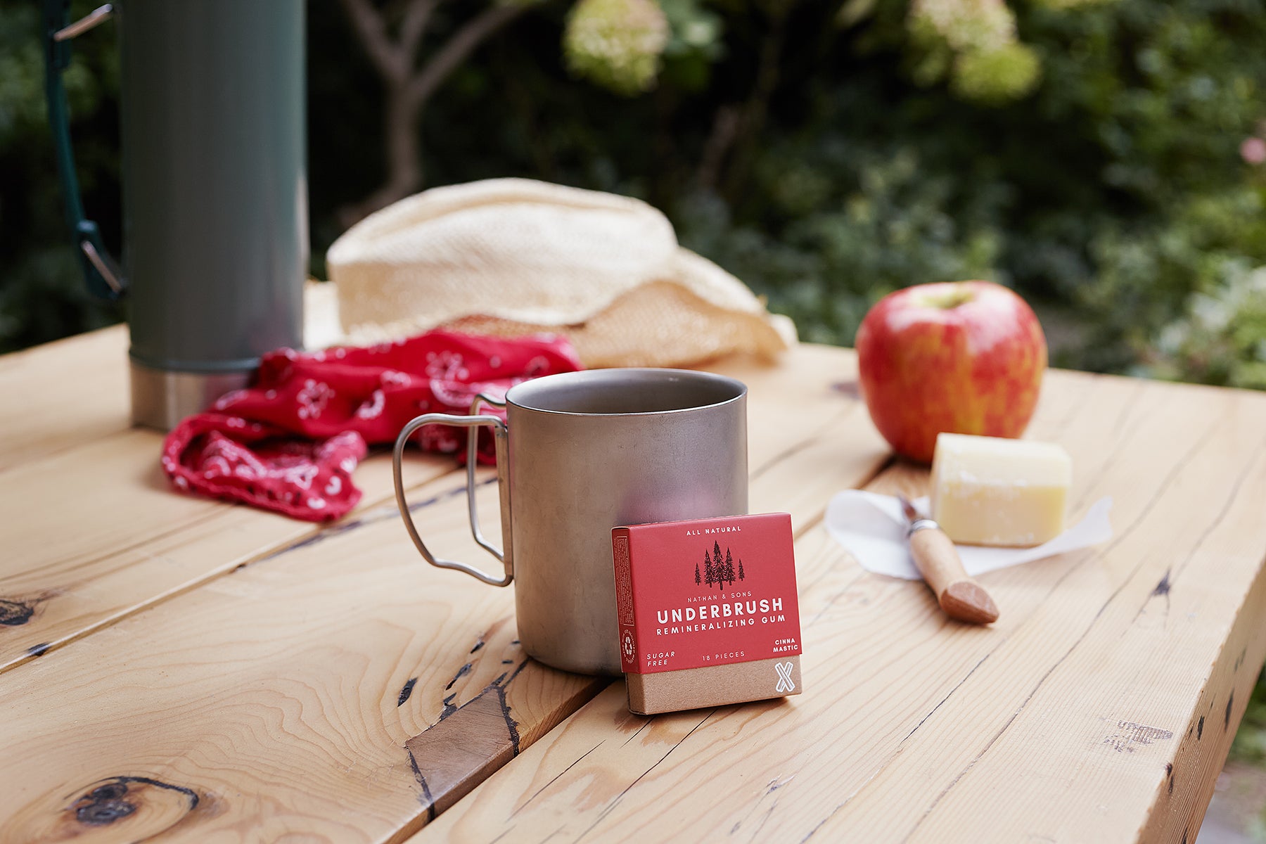 Underbrush cinna mastic remineralizing gum on a rustic outdoor table beside metal mug, apple, and cheese