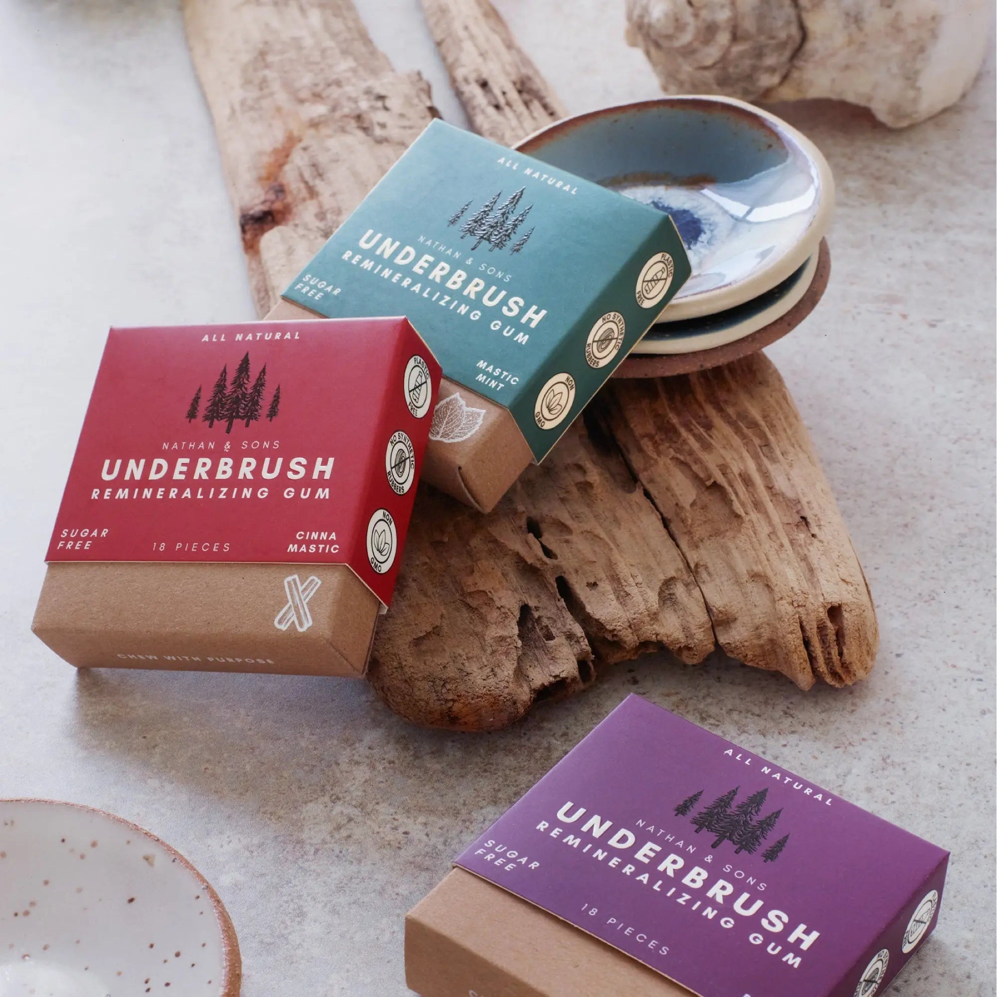 Three boxes of Underbrush Remineralizing Gum in Cinna Mastic, Mastic Mint, and an Berry flavor, displayed on rustic wood with stacked ceramic gum dishes in the background.