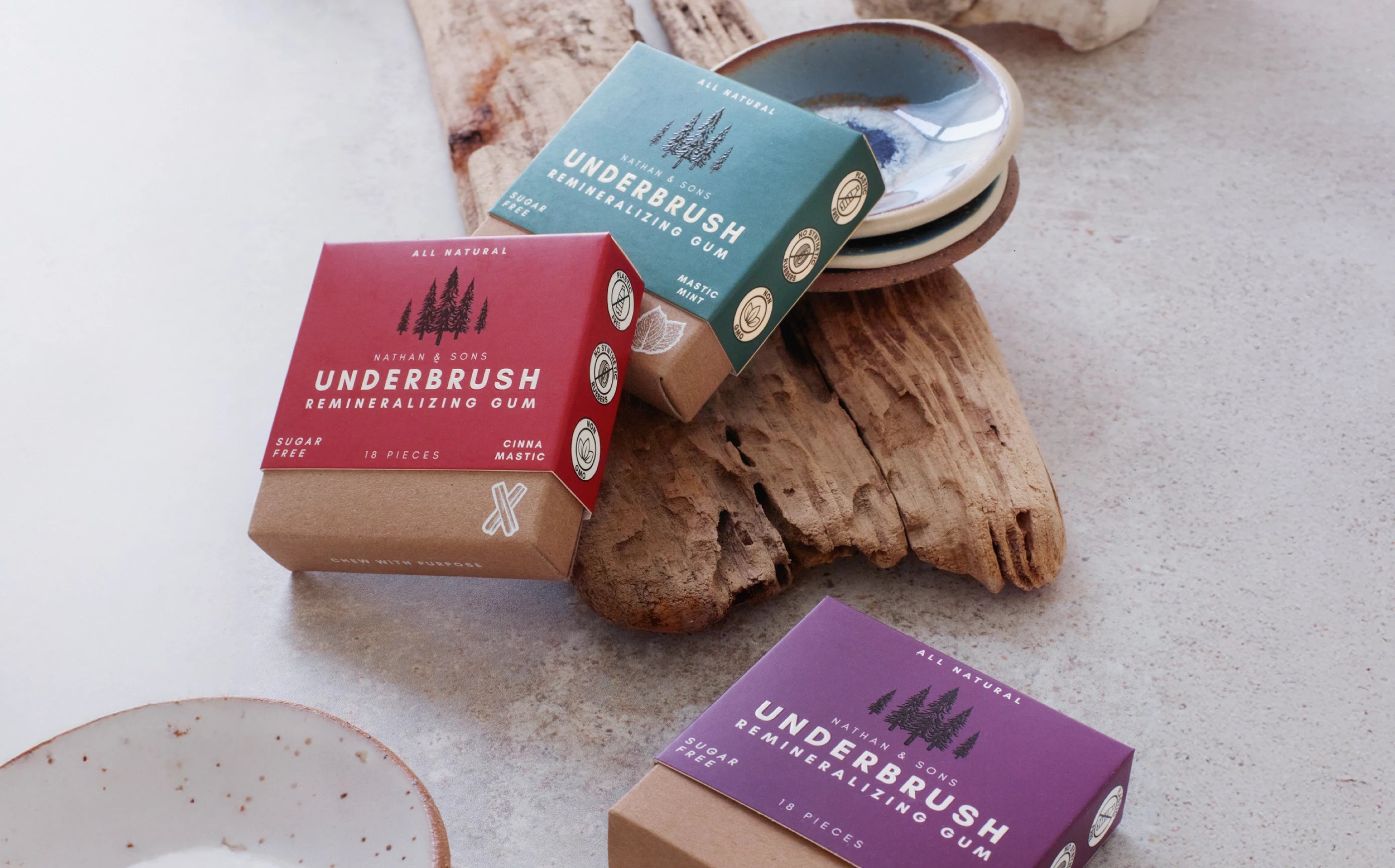Underbrush Remineralizing Gum in Cinna-Mastic, Mastic Mint, and Berry flavors displayed on natural driftwood and a light stone surface