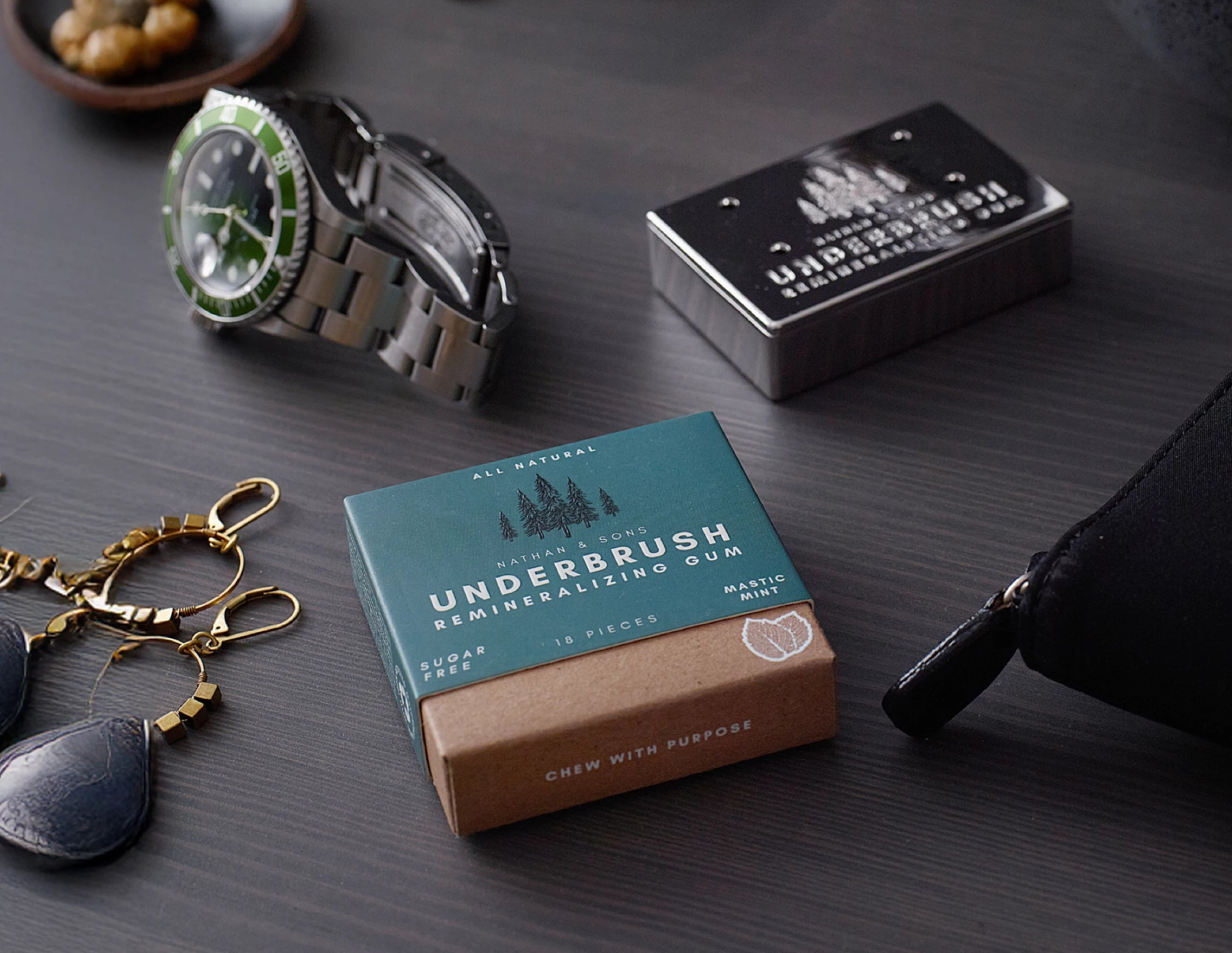 Underbrush Remineralizing Gum in Mastic Mint flavor placed on a dark wood surface alongside sunglasses, watch, and Nathan and Sons Stainless Slide Snap Gum Case