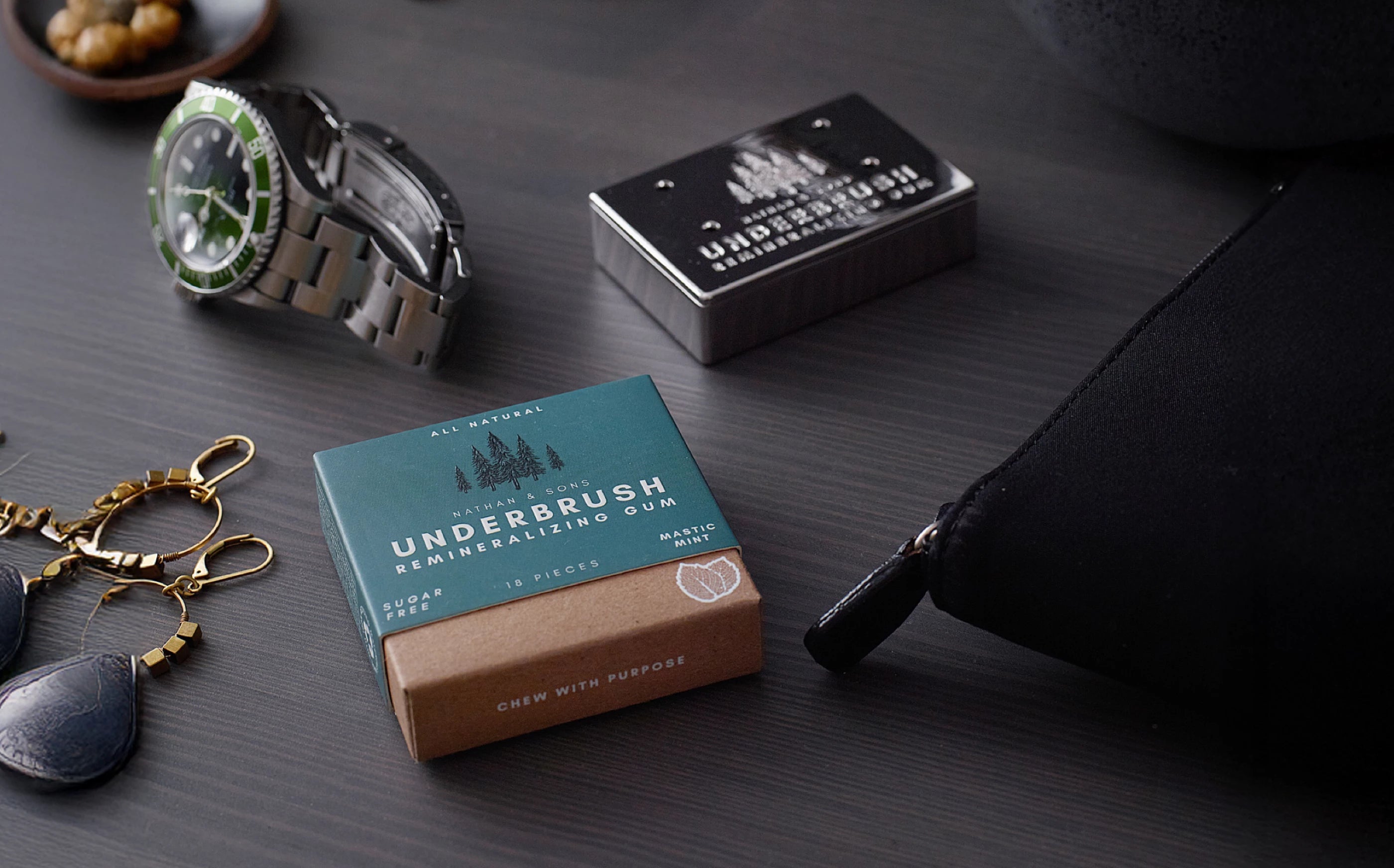 Underbrush Remineralizing Gum in Mastic Mint flavor placed on a dark wood surface alongside sunglasses, watch, and Nathan and Sons Stainless Slide Snap Gum Case