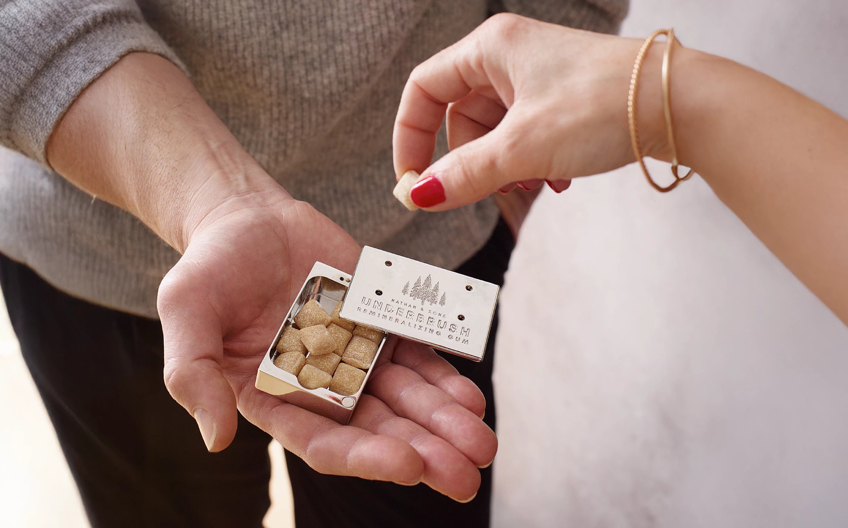 Hand taking Underbrush gum cube from  Stainless Slide Snap Gum Case that is offered by another hand