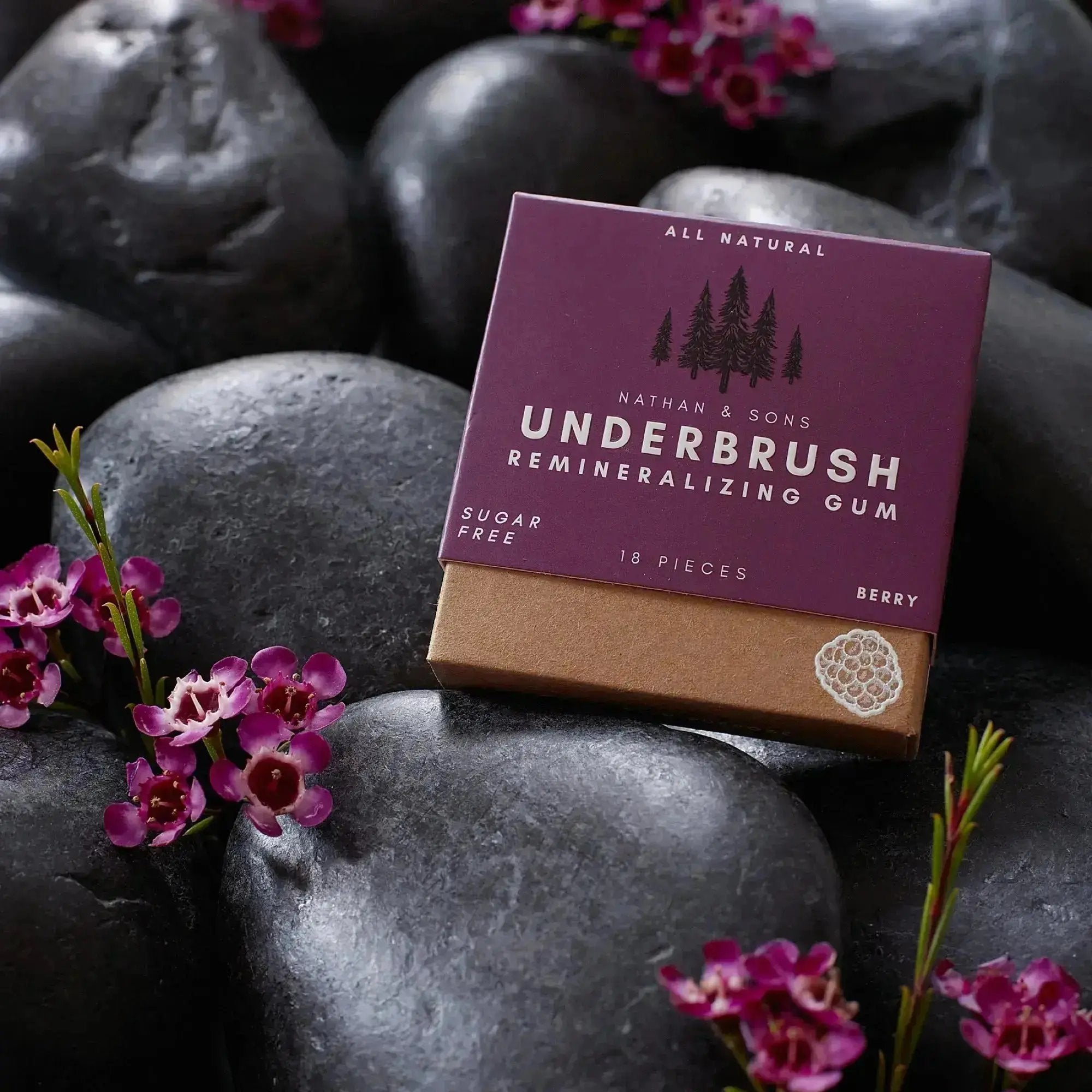 Close-up of a box of Nathan & Sons Underbrush Remineralizing Gum in berry flavor, placed on a surface decorated with small stones and purple flowers