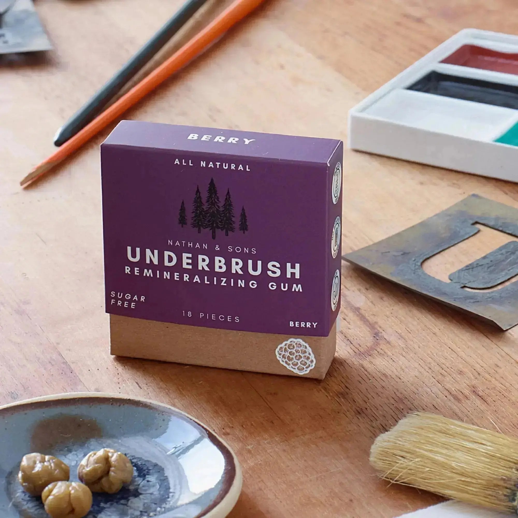 Box of Nathan & Sons Underbrush Remineralizing Gum in berry flavor, placed on a wooden table surrounded by art supplies, including paintbrushes, a metal stencil, watercolor paints, and a small ceramic plate with three chewed Underbrush gum pieces