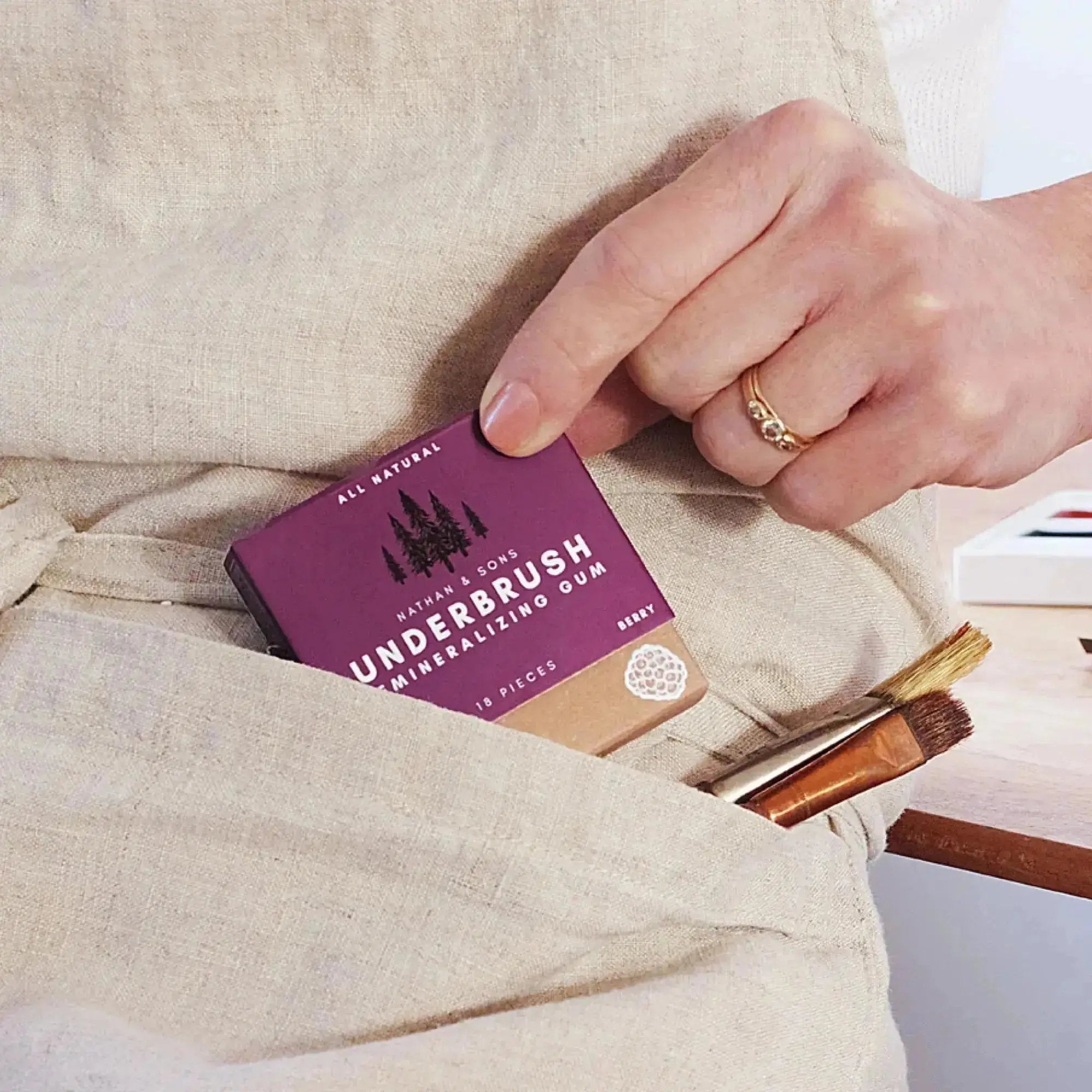 Artist tucking Underbrush berry flavored gum into apron pocket, with paintbrushes and watercolor palette on wooden desk