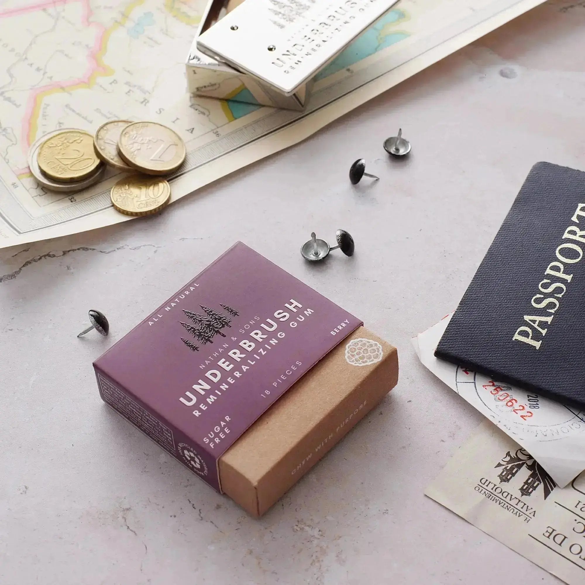 Underbrush Remineralizing Gum in Berry flavor placed beside a passport, European coins, push pins, and travel documents