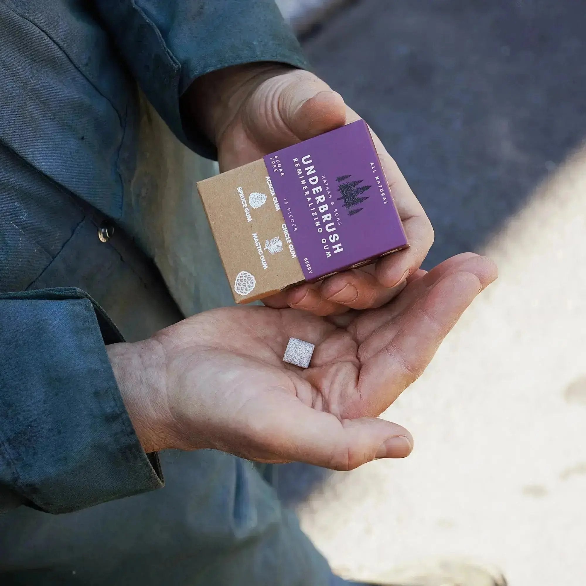 Person wearing a green jacket holding a box of Nathan & Sons Underbrush Remineralizing Gum Berry flavor in one hand, with a single gum piece resting in the other hand.