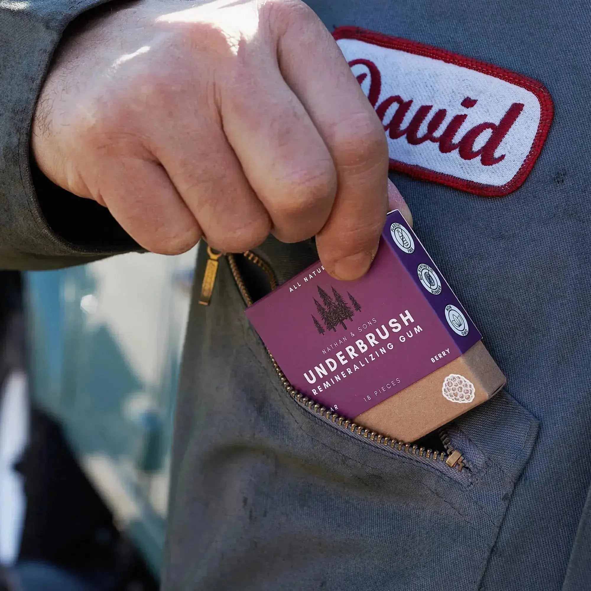 Person wearing a work uniform with a name patch reading "David" placing a box of Nathan & Sons Underbrush Remineralizing Gum, berry flavor, into a zippered chest pocket