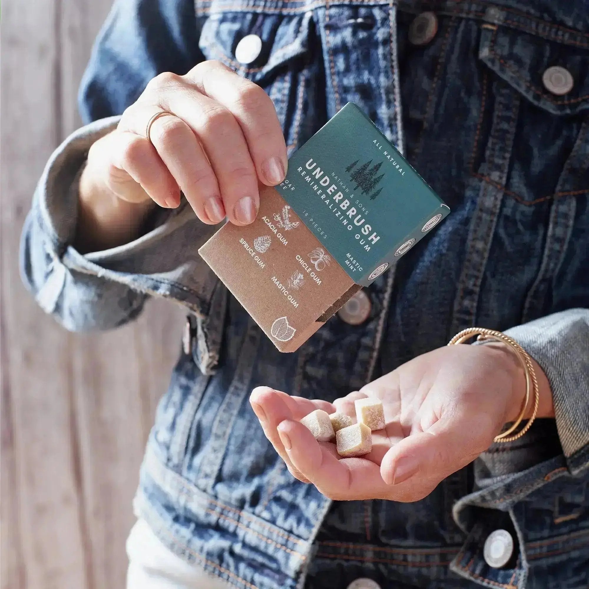 Person wearing a denim jacket pouring small cube-shaped pieces of Nathan & Sons Underbrush Remineralizing Gum Mastic Mint flavor from box into their hand