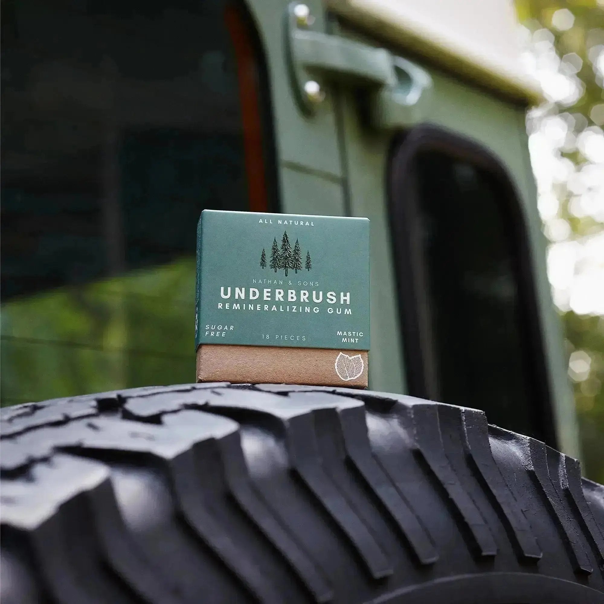 Nathan & Sons Underbrush Remineralizing Gum Mastic Mint flavor box resting on the tread of a large off-road vehicle tire, with part of a green vehicle in the background.