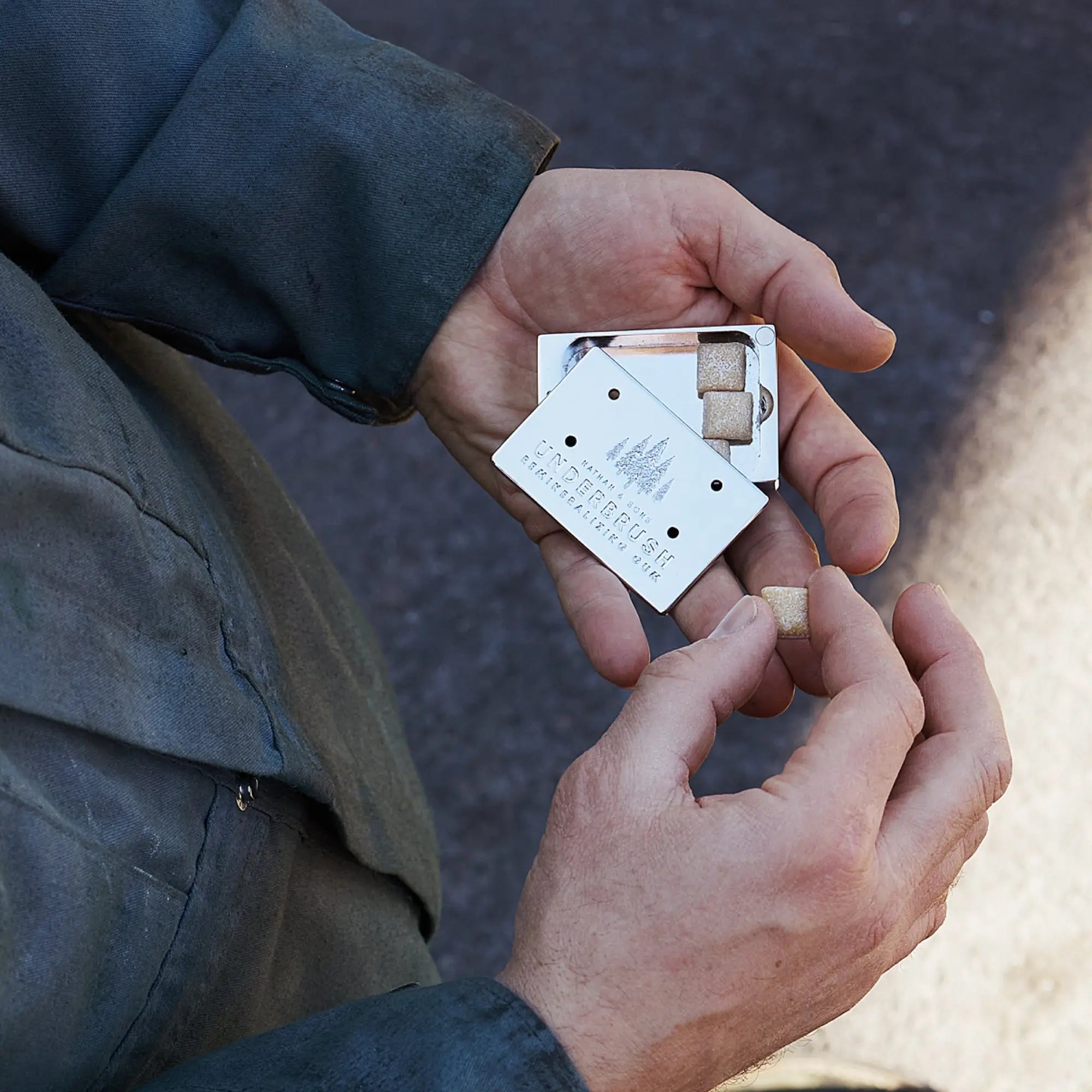 A rugged hand holds the Stainless Slide Snap Gum Case featuring the UNDERBRUSH logo and engraved pine tree design. The lid is partially open, showcasing the Underbrush mineralizing gum on the inside