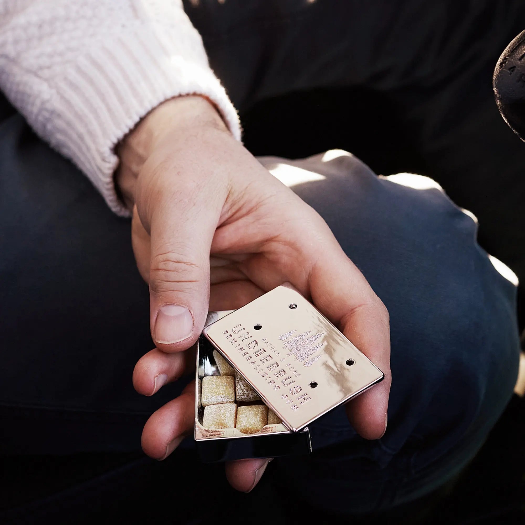 Hand holding Stainless Slide Snap Gum Case featuring the UNDERBRUSH logo and engraved pine tree design. The lid is partially open, showcasing Underbrush mineralizing gum on the inside