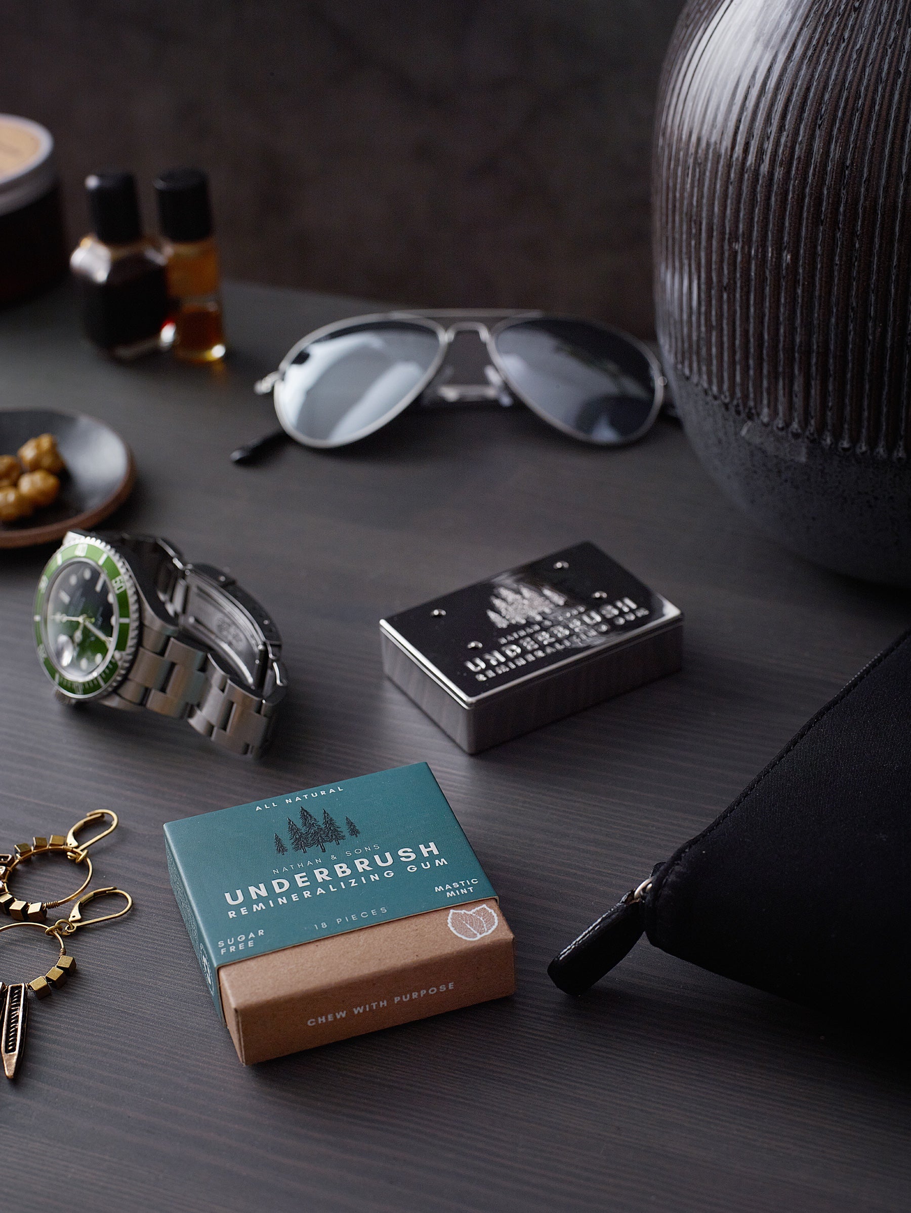 Underbrush Remineralizing Gum in Mastic Mint flavor placed on a dark wood surface alongside sunglasses, watch, and Nathan and Sons Stainless Slide Snap Gum Case