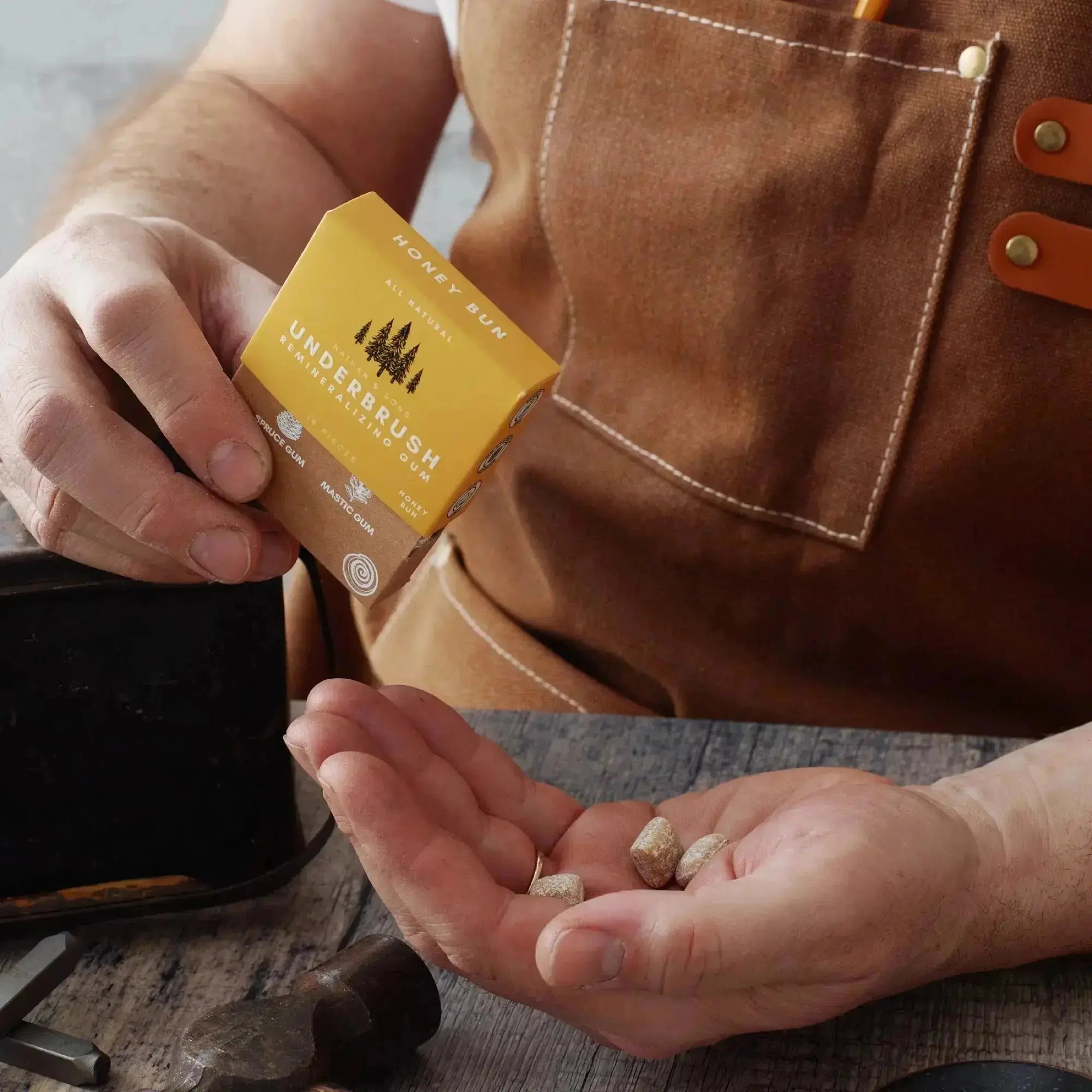 Person wearing a brown apron holding a box of Nathan & Sons Underbrush Remineralizing Gum Honey Bun flavor in one hand, with gum pieces resting in the other hand.