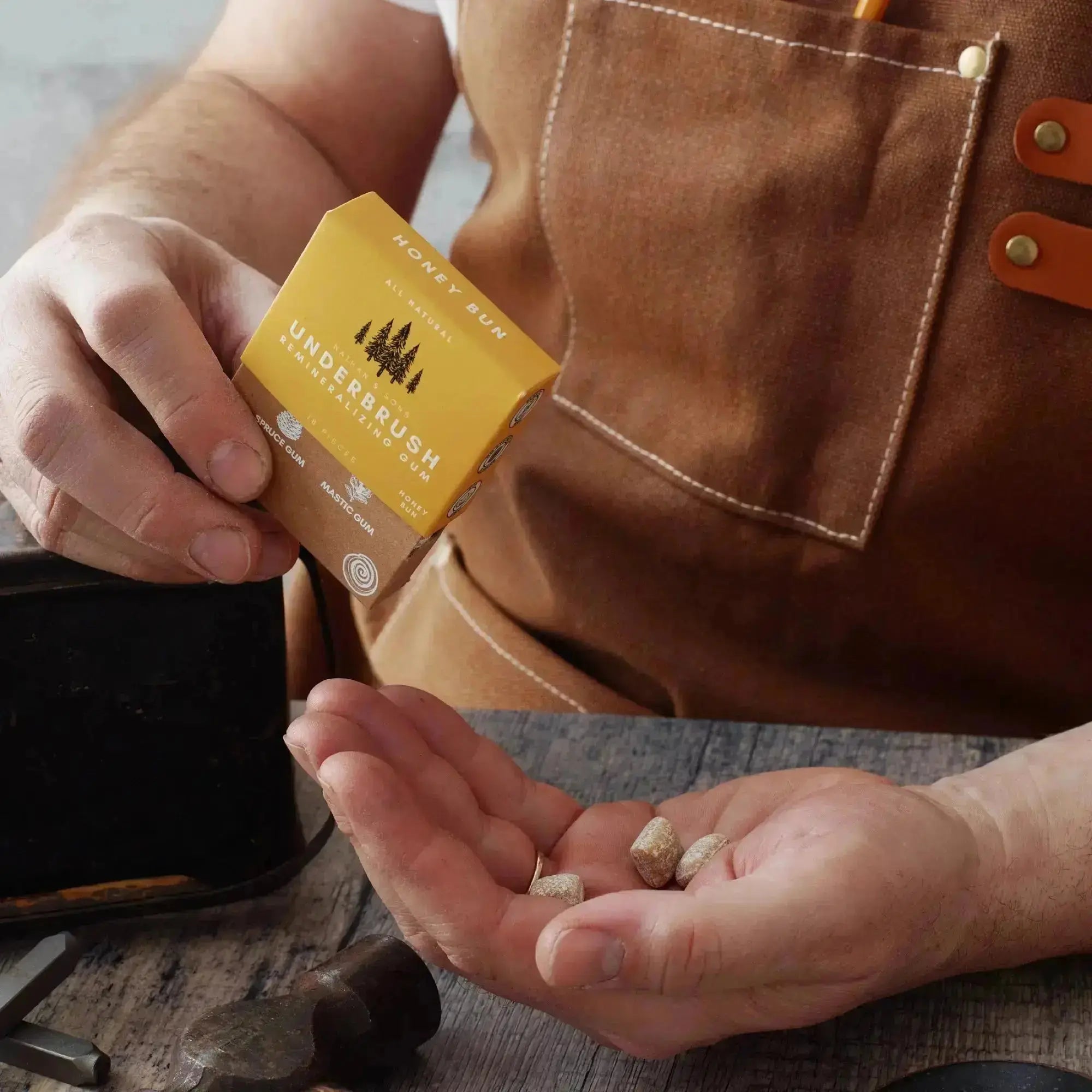 Person wearing a brown apron holding a box of Nathan & Sons Underbrush Remineralizing Gum Honey Bun flavor in one hand, with gum pieces resting in the other hand.