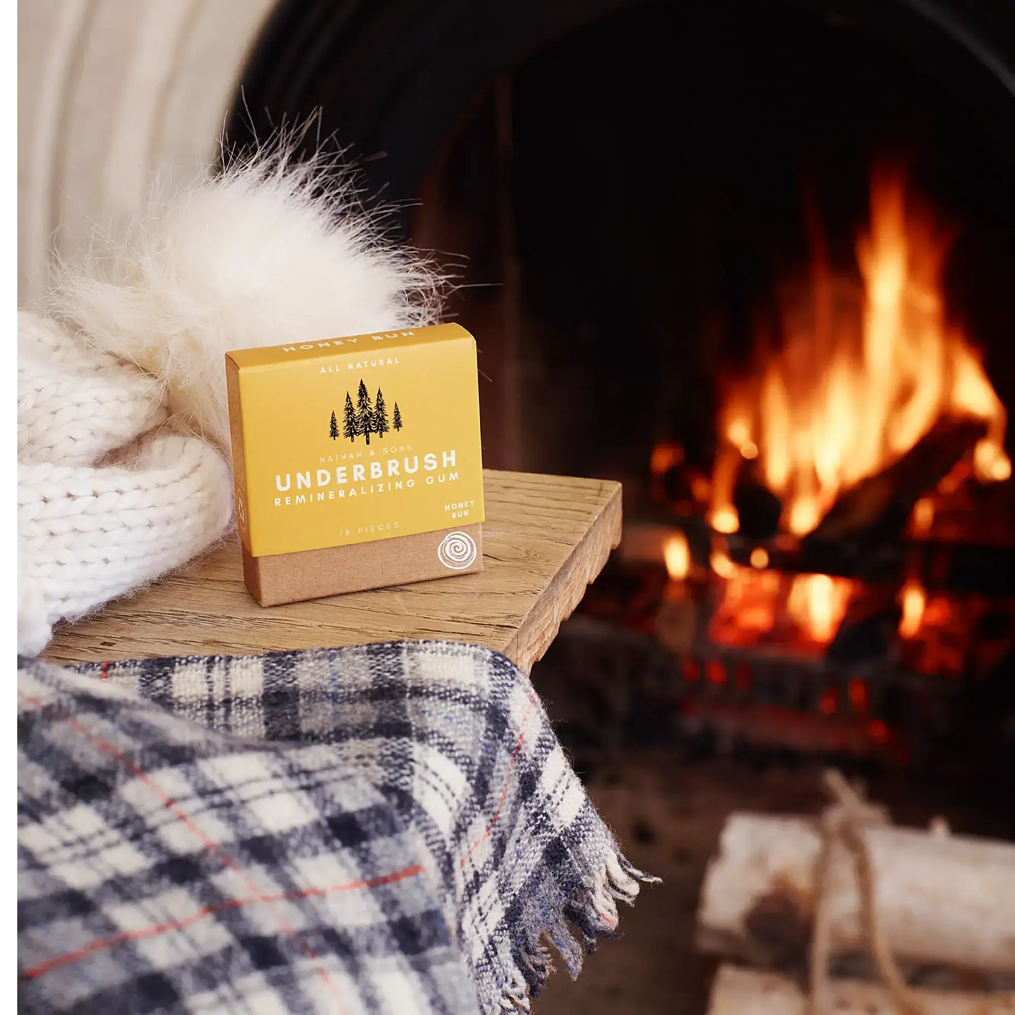 Box of Nathan & Sons Underbrush Remineralizing Gum Honey Bun flavor resting on a wooden table beside a soft white knitted hat and a plaid scarf with a fireplace in the background.