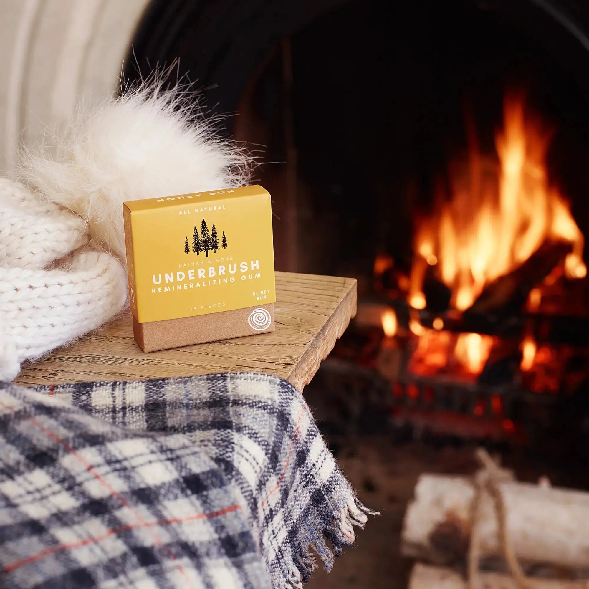 Box of Nathan & Sons Underbrush Remineralizing Gum Honey Bun flavor resting on a wooden table beside a soft white knitted hat and a plaid scarf with a fireplace in the background.
