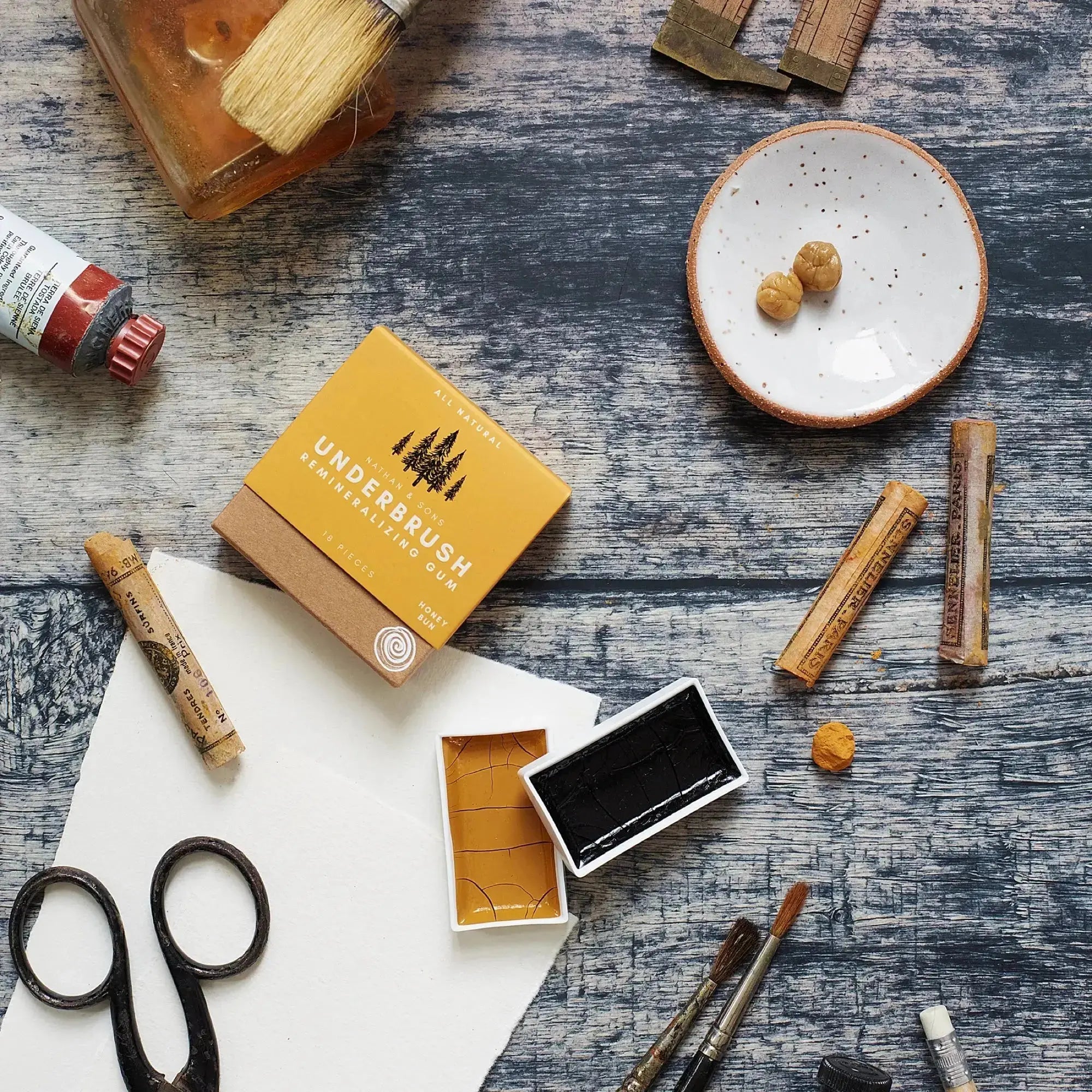 Box of Nathan & Sons UnderbrushRemineralizing Gum Honey Bun flavor surrounded by paintbrushes, watercolor pans, scissors, paint tubes, and a Nathan and Sons Speckled White hand made gum dish with two chewed gums.