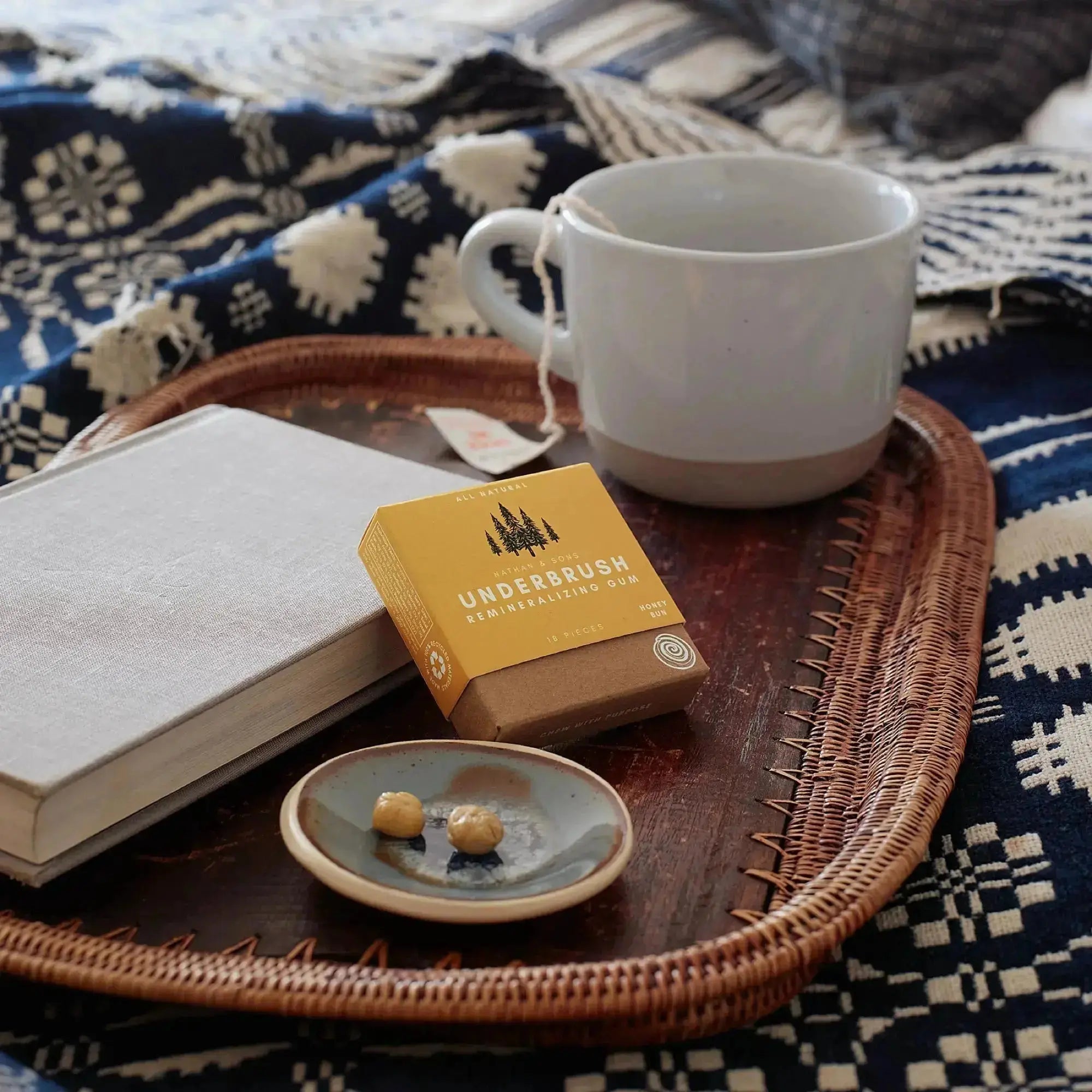 box of Nathan & Sons Underbrush Remineralizing Gum Honey Bun flavor)placed on a woven tray alongside a white mug of tea, a closed book, and a Nathan and Sons Batik Blue handmade gum dish with two chewed gum pieces.