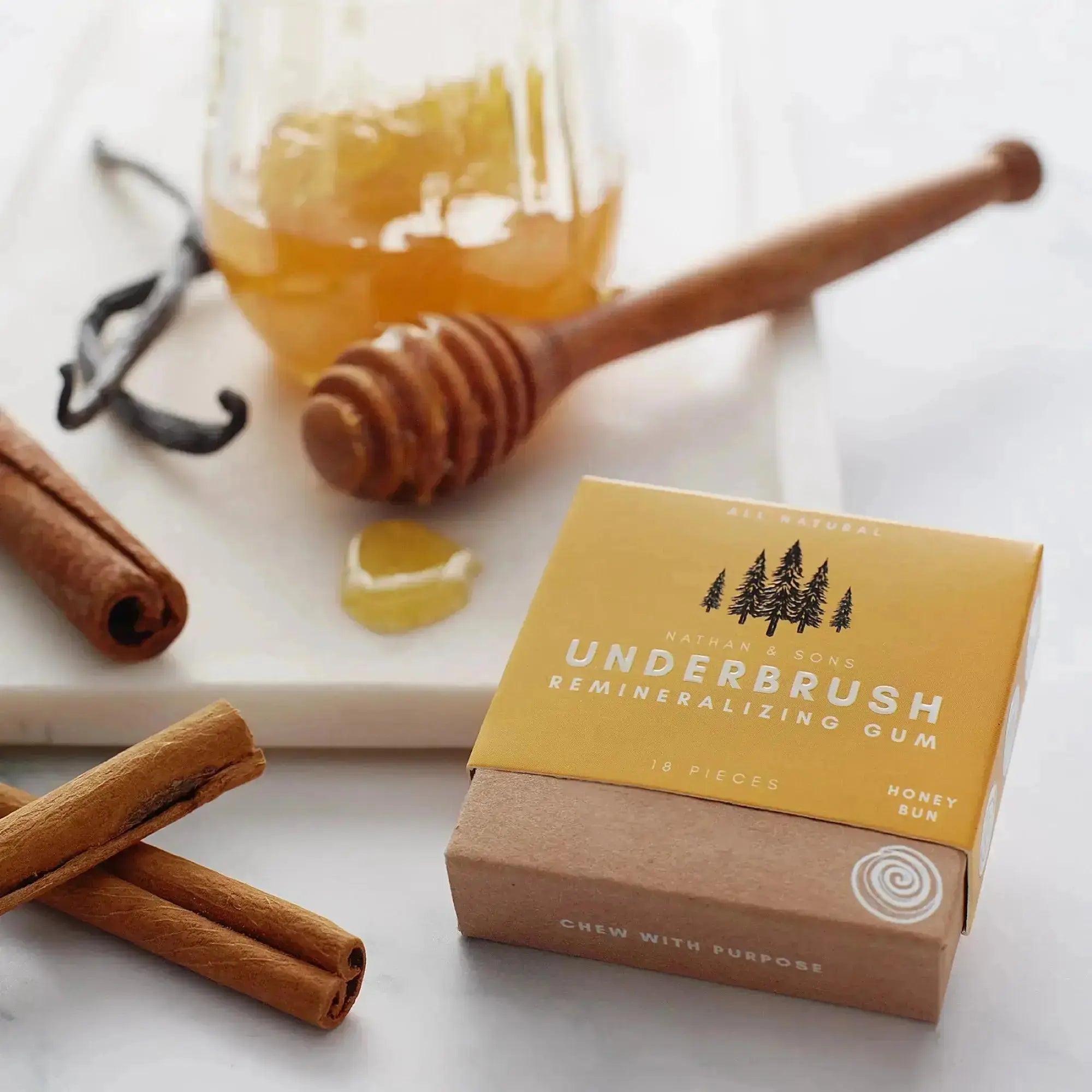 Box of Nathan & Sons Underbrush Remineralizing Gum Honey Bun flavor placed on a marble surface beside cinnamon sticks, a honey dipper with dripping honey, a jar of honey.