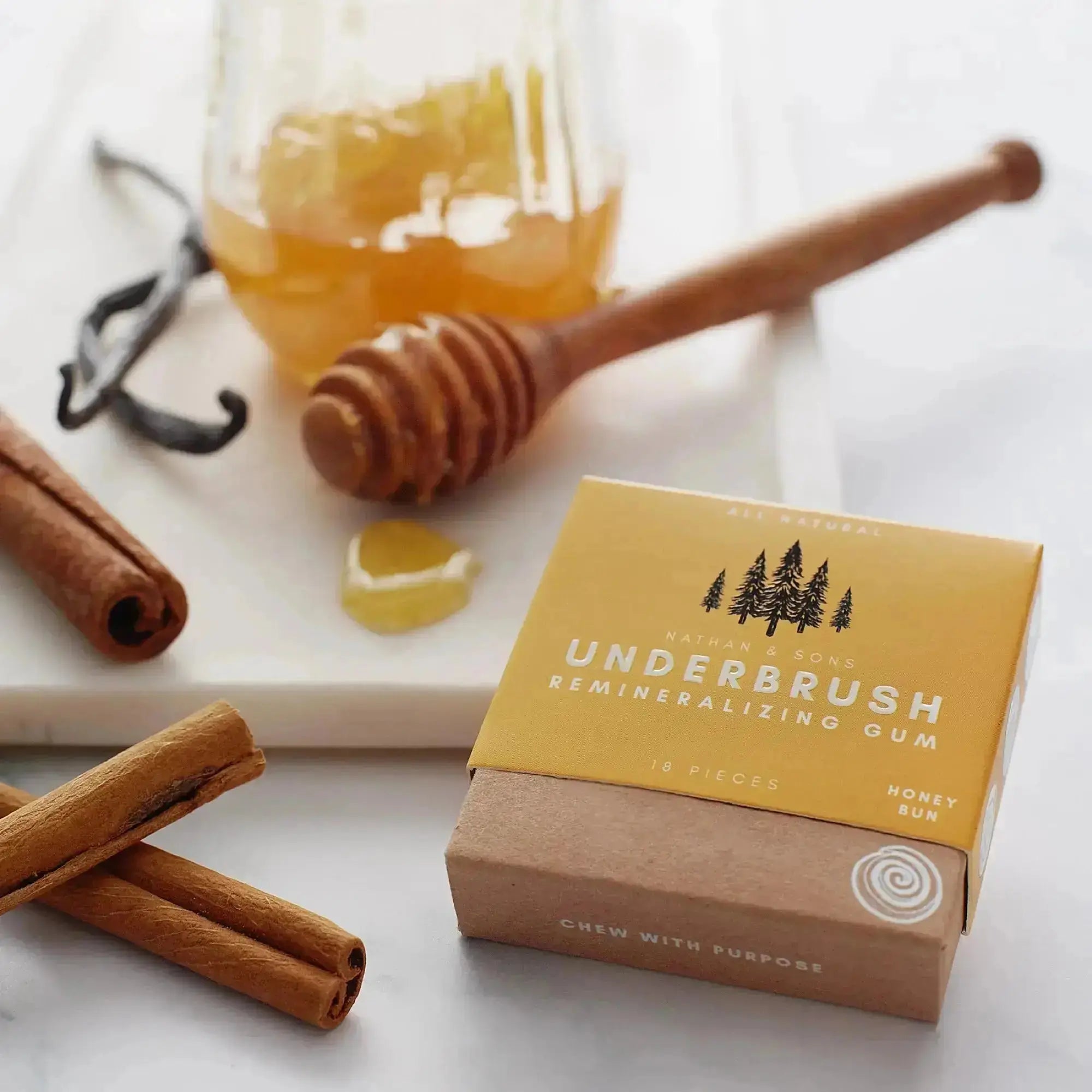 Box of Nathan & Sons Underbrush Remineralizing Gum Honey Bun flavor placed on a marble surface beside cinnamon sticks, a honey dipper with dripping honey, a jar of honey.