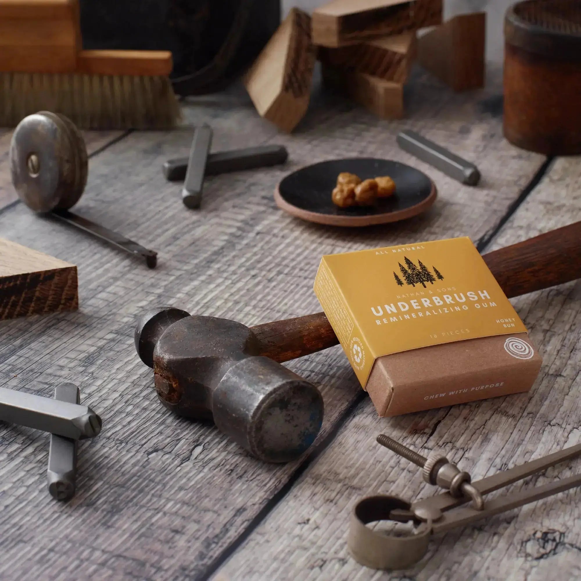 Box of Nathan & Sons Underbrush Remineralizing Gum Honey Bun flavor resting on a weathered hammer surrounded by vintage woodworking tools, metal stamps, and wooden blocks on a rustic wooden surface.